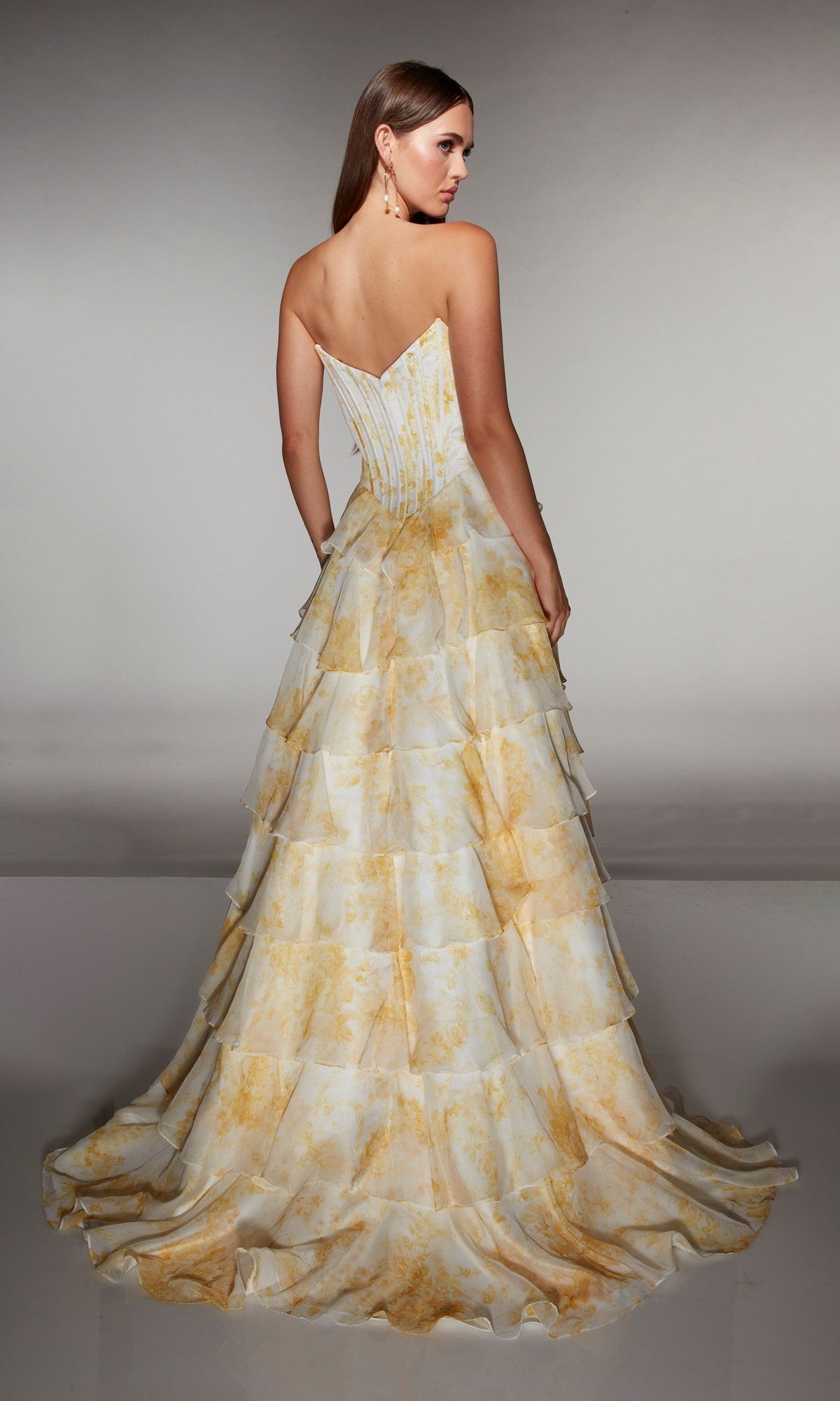 A woman is shown from the back wearing the Alyce 62192 long prom dress, featuring a strapless fitted corset bodice and a tiered yellow-patterned chiffon ruffle skirt.